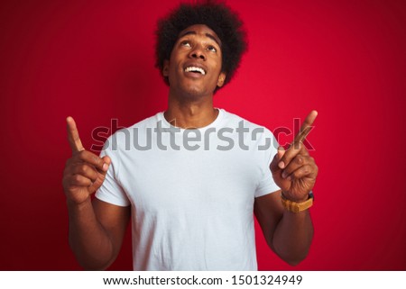 Young american man with afro hair wearing white t-shirt standing over isolated red background amazed and surprised looking up and pointing with fingers and raised arms.