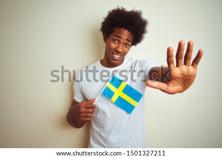 Young afro american man holding Sweden Swedish flag standing over isolated white background with open hand doing stop sign with serious and confident expression, defense gesture