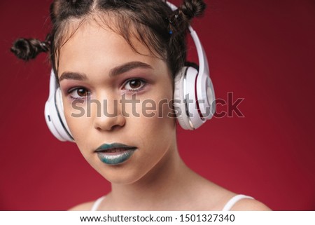 Image closeup of teenage punk girl with bizarre hairstyle and dark lipstick listening to music with headphones isolated over red background