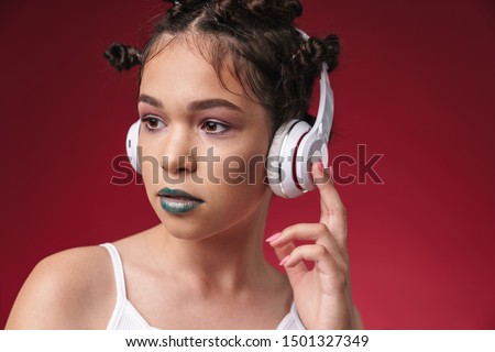 Image closeup of lovely punk girl with bizarre hairstyle and dark lipstick listening to music with headphones isolated over red background