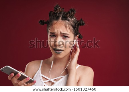 Image of displeased punk girl with bizarre hairstyle and dark lipstick frowning while using smartphone with earphones isolated over red background