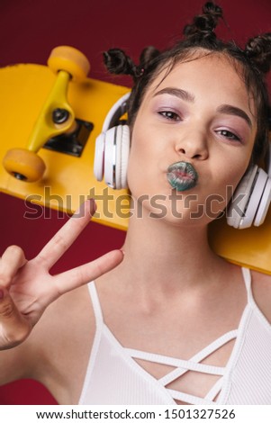 Image of stylish punk girl with bizarre hairstyle and dark lipstick holding skateboard while listening to music with headphones isolated over red background