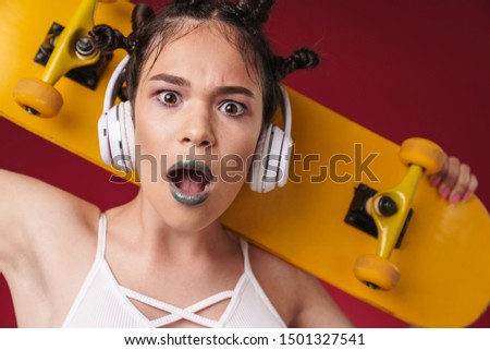 Image of surprised punk girl with bizarre hairstyle and dark lipstick holding skateboard while listening to music with headphones isolated over red background