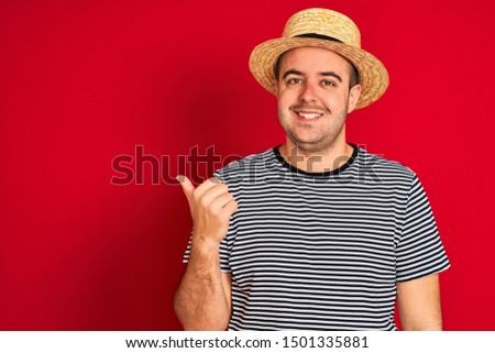 Young man wearing striped navy t-shirt and hat standing over isolated red background smiling with happy face looking and pointing to the side with thumb up.