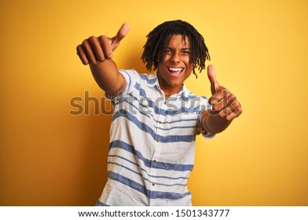 Afro american man with dreadlocks wearing striped shirt over isolated yellow background approving doing positive gesture with hand, thumbs up smiling and happy for success. Winner gesture.