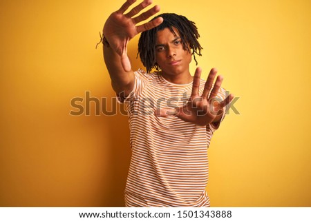Afro man with dreadlocks wearing striped t-shirt standing over isolated yellow background doing frame using hands palms and fingers, camera perspective