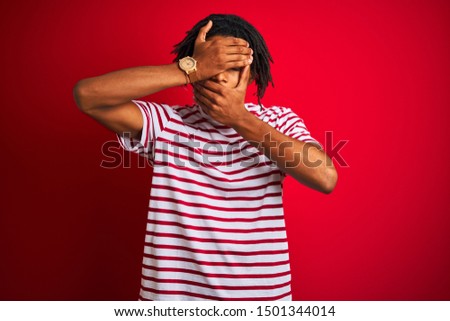 Young afro man with dreadlocks wearing striped t-shirt standing over isolated red background Covering eyes and mouth with hands, surprised and shocked. Hiding emotion