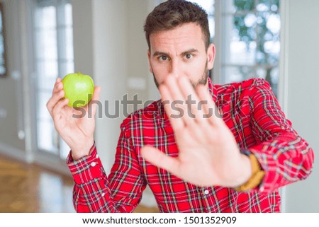 Handsome man eating fresh healthy green apple with open hand doing stop sign with serious and confident expression, defense gesture