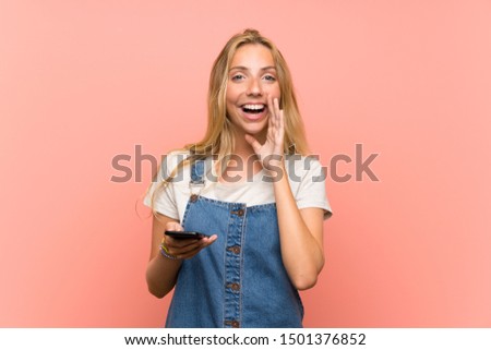 Blonde young woman with a mobile phone over isolated pink wall with surprise and shocked facial expression