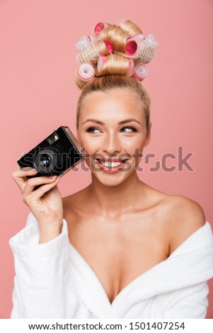 Beautiful smiling young girl wearing bathrobe and curlers standing isolated over pink background, holding photo camera
