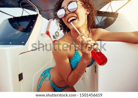 Laughing young woman wearing sunglasses and a bikini sipping on a drink while standing on a boat during summer vacation