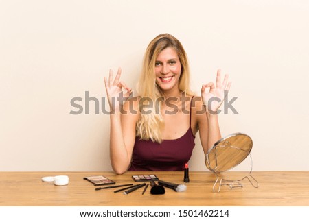 Young blonde woman with cosmetic in a table showing an ok sign with fingers