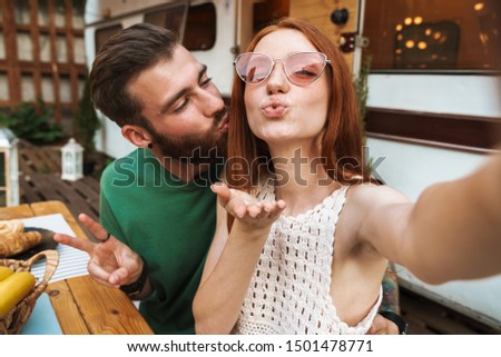 Happy smiling couple having lunch while sitting at the table outdoors, camping at the trailer, taking a selfie, sending kiss