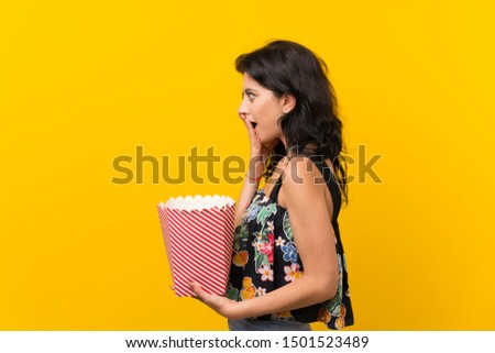 Young woman over isolated yellow background holding a bowl of popcorns