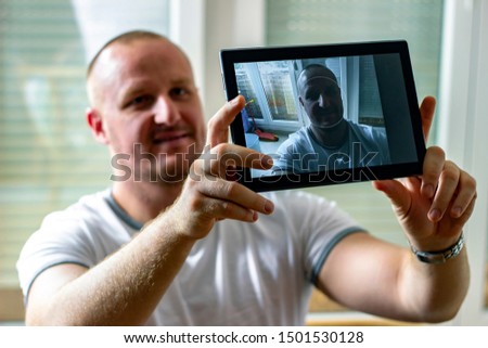 Young man standing and taking selfie with his tablet. Taking pictures for his love. Smiling young man in white t shirt making selfie at home while smiling at camera. Tehnology concept. Copy-space. 