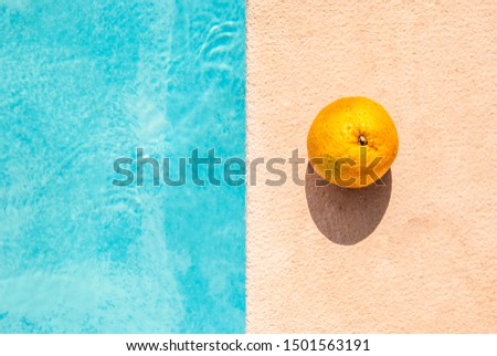 An orange fruit lies on the sandstone texture side of the pool with blue water. Hard light and contrast shadow. Minimal still life.