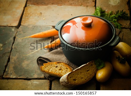 Closed casserole dish with fresh carrots and potatoes and slices of crusty bread on weathered bricks with copyspace