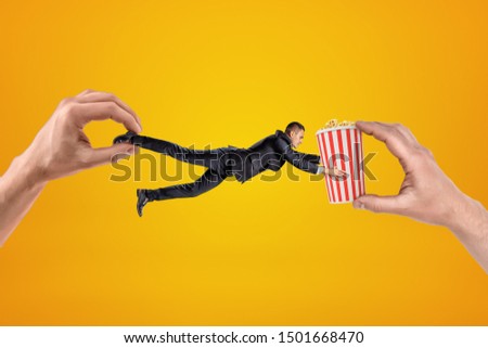 Big male hand holding tiny businessman who is reaching to another big hand holding popcorn bucket on yellow background. People and objects. Digital art. Cinema industry.