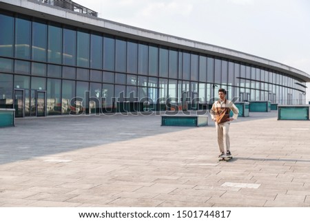 Afro-american young man in tracksuit standing on skateboard ready to practice making stunts outdoors in business area near glass office building at sunny day. Extreme sports, active lifestyle, hobby