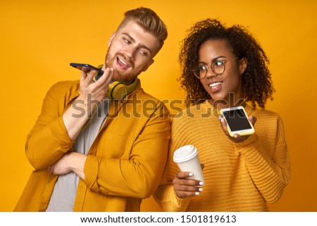 Happy black woman and ginger man keep modern cell phones near mouthes, make a voice call, enjoy nice conversation, drink hot beverage from a disposable cup, stand back to each other over orange wall