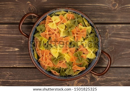 Lot of whole red, yellow and green uncooked farfalle in old pot flatlay on brown wood