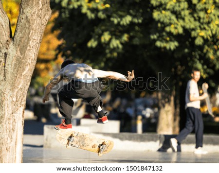 A young man in a city park makes a skateboard jump. Back view with blurry background.