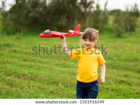A preschooler girl in a yellow T-shirt and jeans holds over her head and launches a red toy airplane to the left on the street. autumn evening. horizontal photograph of a child.