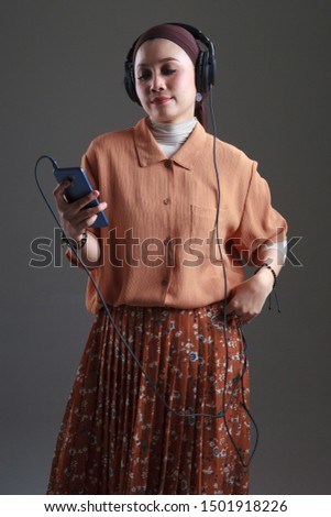 Beautiful asian muslim in headphones listens to music and dancing using smartphone isolated grey background.