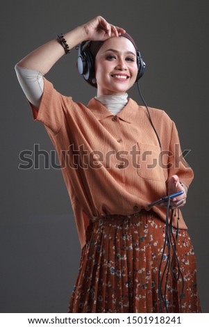 Beautiful asian muslim in headphones listens to music and dancing using smartphone isolated grey background.