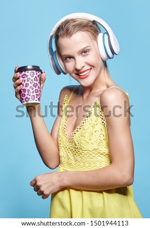 Young pretty happy woman drinking coffee and listening to music in headphones. Female in yellow dress on blue background. Smiling girl with coffee paper cup in hand and big earphones