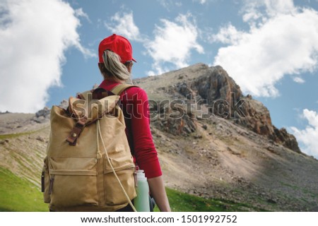 View from the back Girl athlete in a red cap sunglasses and a yellow backpack stands on a green slope against the background of the epic cliffs of the Caucasus