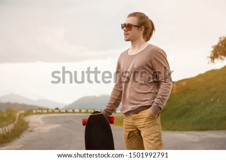 Stylish young man standing along a winding mountain road with a skate or longboard on his shoulder in the evening after sunset. The concept of youth sports and travel hobbies