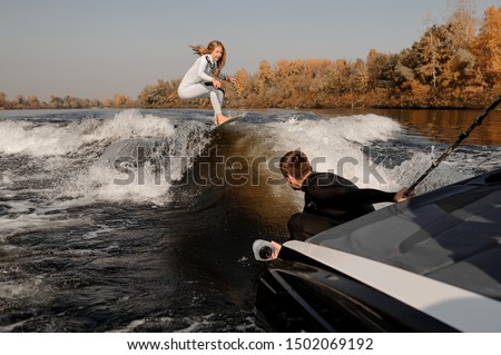 Photographer taking pictures with a camera in moisture proof cover from the motorboat holding the rope of the girl riding on the wake surf