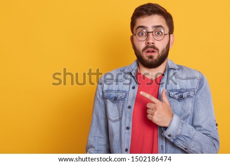 Image of shocked young man pointing finger aside at blank copy space, astonished surprised guy dressescasual clothing, looking directly at camera, standing isolated over yellow studio background.