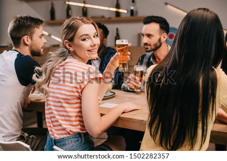 selective focus of cheerful young woman smiling at camera while holding glass of light beer