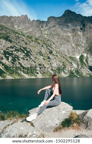 Young beautiful sporty girl in grey pants sitting on the big rock and posing on the background of mountain lake, active lifestyle concept, summer holidays