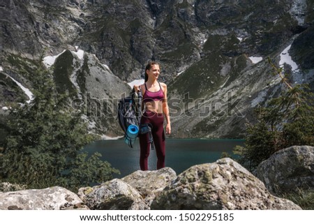 Young brunette girl traveler in red sportswear with backpack in hand standing on the stones, amazing mountine lake on the background, active and healthy lifestyle concept, summer time