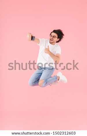 Image of cheerful man wearing eyeglasses gesturing thumb up and taking selfie on cellphone while jumping isolated over pink background