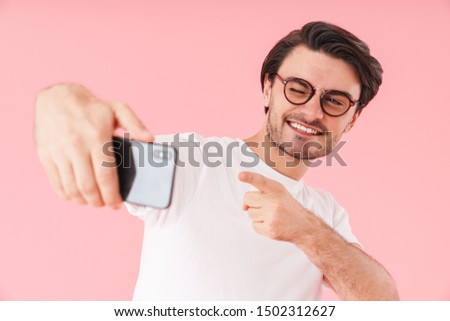 Image of joyful man wearing eyeglasses pointing finger at camera and taking selfie photo on cellphone isolated over pink background