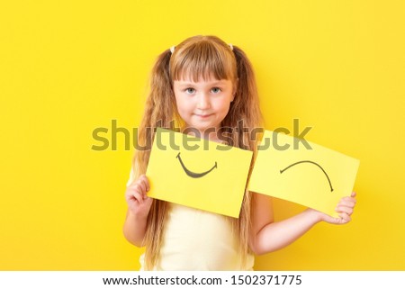 Little girl with drawn smiling and upset mouths on sheets of paper against color background