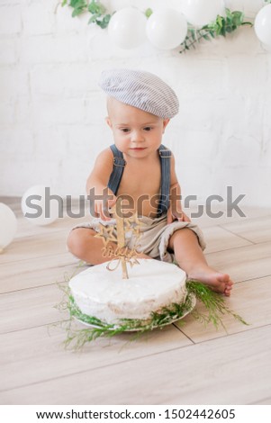 Little cute boy in suspenders and vintage cap. First birthday with a birthday cake and decor with white balloons and sprigs of eucalyptus. Rustic First Birthday Party
