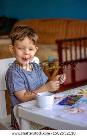 Toddler making funny face,having fun at home,while painting on white board. Little boy with hands painted in colorful paints ready for hand prints. Cute little child boy with colored hands. Copy-space