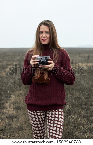 Young female wearing stylish sweater and 
leggings with a pattern hold old vintage film camera outdoors in the wild plain field at autumn foggy cold weather