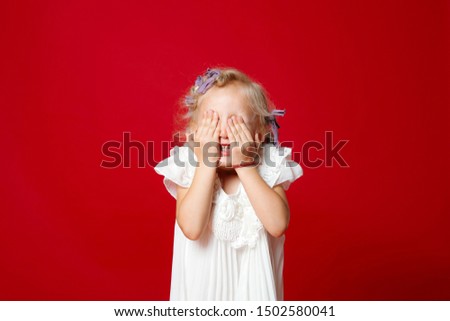 Happy young blonde curly model girl hiding her face with hands in white dress isolated on red. Little girl playing hide and seek
