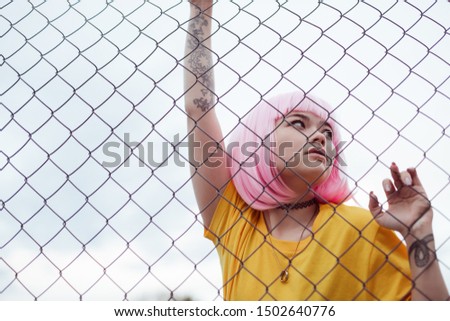 Low angle of Asian teen girl in pink wig and stylish outfit grasping net fence and looking away while spending time on modern city street Royalty-Free Stock Photo #1502640776