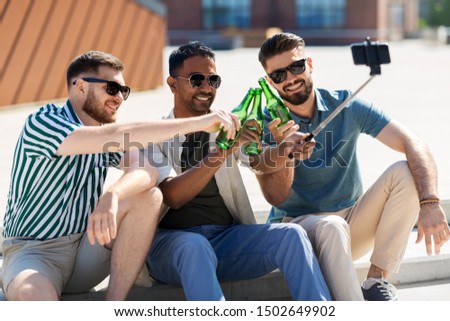 leisure, technology and people concept - happy male friends taking picture by smartphone on selfie stick and toasting beer on street in summer