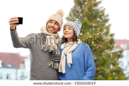 people, technology and winter holidays concept - happy couple in knitted hats and scarves taking selfie by smartphone over christmas tree in old town of tallinn city background