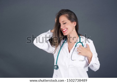 Shot of happy young doctor woman with positive smile, has long hair, rejoices having weekend and good rest after hard working exhausting week, isolated on gray wall.