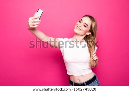 Portrait of a joyful pretty girl listening music with earphones while standing and taking a selfie with mobile phone isolated over pink background