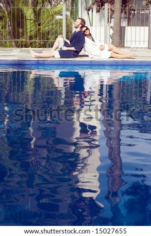 A man and a woman are relaxing together beside a pool.  They are leaning against each other.  The man is looking away from the camera and the woman is looking at it.  Vertically framed photo.